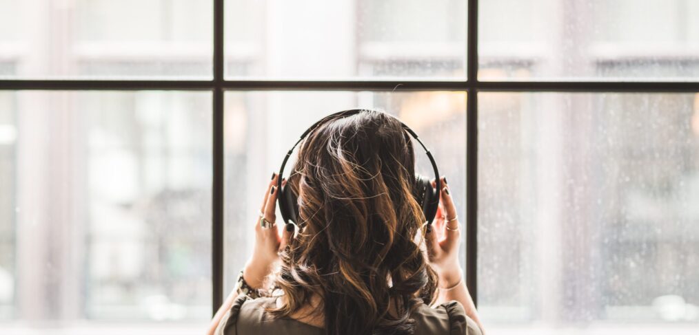girl standing with back to camera, facing a window with headphones on